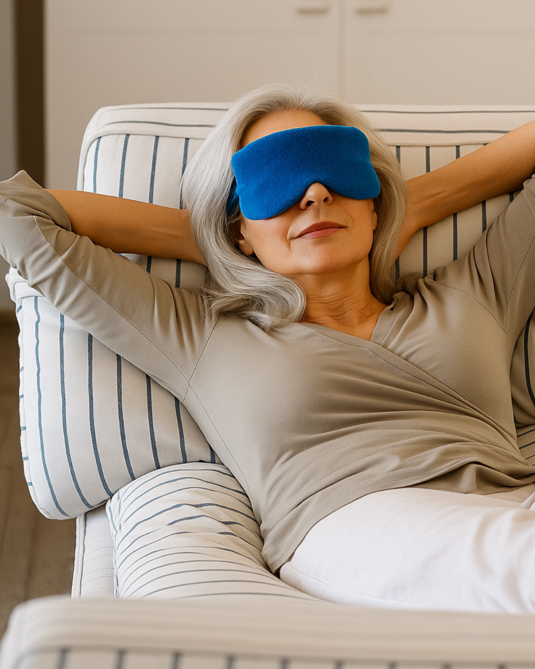 "A woman with gray hair wearing a light brown top and white pants relaxes on a striped chair with her hands behind her head, using a bright blue sleep mask."